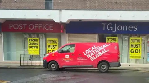 A red, Royal Mail post van is parked on a street outside of a Post Office branch. The windows of the building are partially covered with large yellow posters which reads "store closing down" and "everything must go". A red and white sign above the door reads "Post Office".