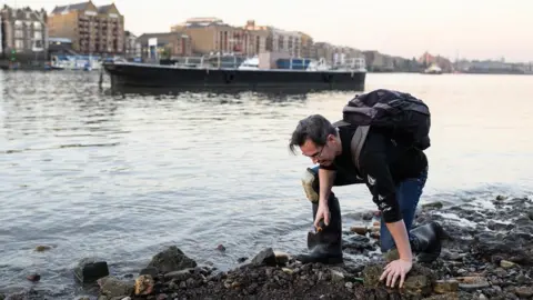Getty Images Mudlark Jason Sandy wearing a backpack is bent over and holding a trowel as he searches the shoreline of the River Thames for historical items