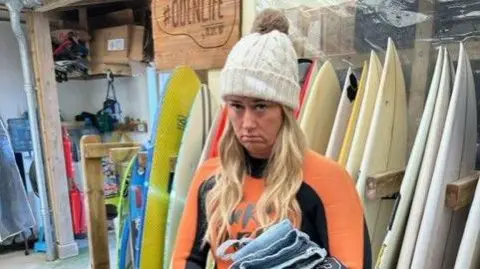 Simon Tite Emma Frances in an orange and black wetsuit on the day of the marriage proposal. Ms Frances has blonde hair and is wearing a cream bobble hat. She is standing in front of surfboards and is not looking very impressed. 