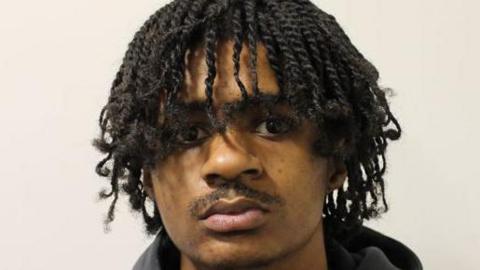 A police mugshot of Kemani Duggan looking directly into the lens. He has got short braids and a moustache and a black collared top.