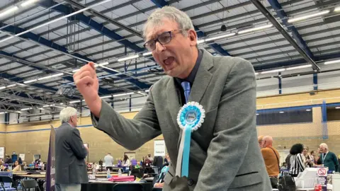 A man wearing a grey blazer and a Reform UK turquoise and white rosette celebrating an election win. A hall with people and tables can be seen in the background.