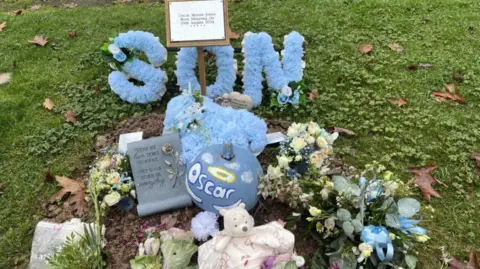 BBC A small grave, is surrounded by grass.  There is a blue wreath that says son in blue flowers and a blue pumpkin that says Oscar with a yellow halo above the name.