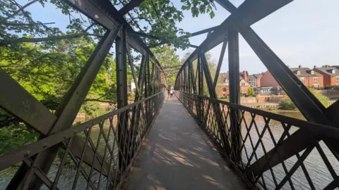 BBC A long bridge walkway is covering a river. It is surrounded by blue sky, houses and green trees. In the long distance, we can see a person standing in the middle of the bridge.