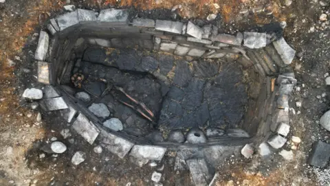 An aerial view of the burial site showing the stone-lined pit with a skeleton tucked into a corner. There is bare earth around the stone lip of the pit.