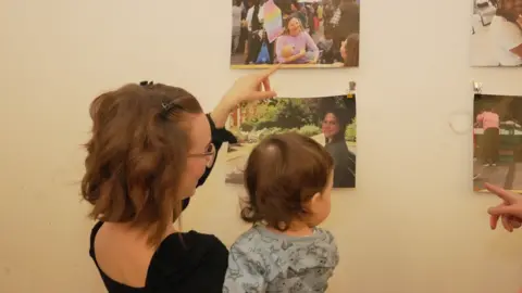 City of Wolverhampton Council A woman with short chestnut brown hair and glasses is holding a baby and pointing at pictures on a white wall. the pictures are of women breastfeeding in public places