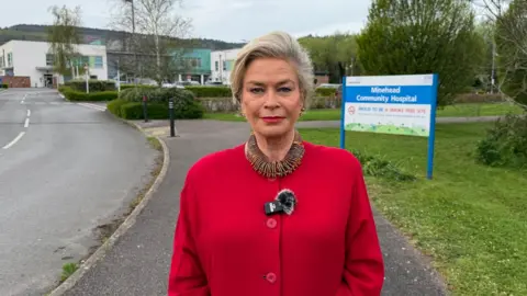 A woman stood in the middle of the frame smiling at the camera, wearing a red coat and extravagant necklace. Behind her the community hospital, and a sign directing people inside.