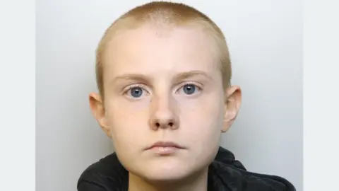 A police mugshot of Alina Burns, wearing a black hooded jacket and a grey T-shirt. She has blue eyes and her blonde hair is closely shaven. She is standing against a light grey background, looking ahead with a blank expression. 