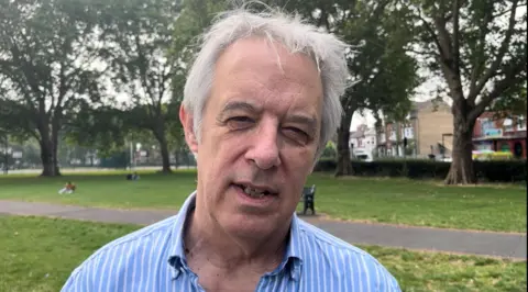 Hugh Flouch standing in a park with trees behind him