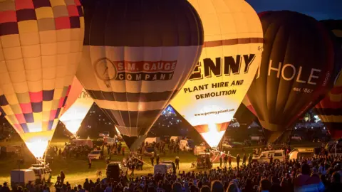 Getty Images Several hot air balloons that are inflated but tethered to the ground. A few of them have fire inside. A large crowd of people can be seen watching the display.