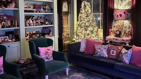 IWC Media Cozy living room decorated for Christmas with a lit tree, colourful pillows on dark sofas, and shelves filled with figurines. A grandfather clock sits next to the Christmas tree.