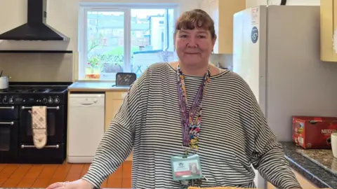 Keeley stands behind her kitchen counter wearing a striped top. She has brown hair, tied back and a lanyard around her neck.