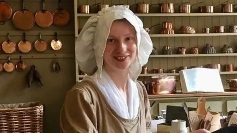 A young woman in a regency costume in a kitchen. She is wearing a beige long sleeved top with a white top underneath, a white apron and a large white hat. 