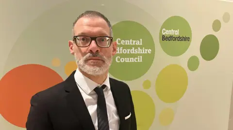 Amy Holmes/BBC Council leader Adam Zerny is dressed in a formal black suit with a white shirt and black tie, standing in front of a display board featuring the Central Bedfordshire Council logo and colourful circular graphics.