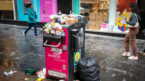 A bin on a Glasgow street, overflowing with rubbish and with people walking by