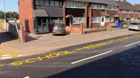 A street with a row of brick houses in the background and School Keep Clear written in yellow paint on the road