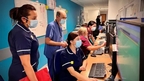 A group of medical staff are standing around a bank of computers in a busy emergency department. They are nearly all wearing face masks.