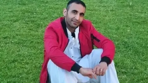 Family photo Abdul Oryakhel looking at the camera. He is sitting on an area of grass and is wearing white clothing with a red jacket on top