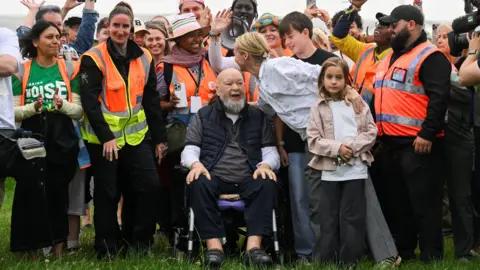 PA Media A line of people, some wearing orange hi-vis vests. In the centre is Michael Eavis, using a wheelchair. He has short white hair and beard and is wearing a black gilet over a grey shirt. A woman in a white blouse is leaning over him. They are all on grass.