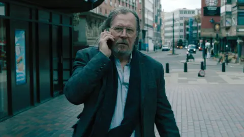 Apple Gary Oldman has long hair and grown out facial hair. He is on his phone walking through a quiet area of a city with buildings surrounding him.