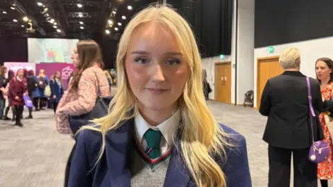Jessica is looking at the camera. She has long blonde hair and is wearing a navy school blazer and a teal coloured tie with a grey jumper.
