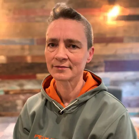 A woman with very short brown hair looks into the camera with a straight face. She wears a grey hoodie with orange inside the hood.