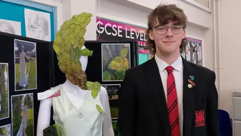 A teenage boy is standing next to a mannequin. He has glasses, brown hair and is wearing his school uniform. The mannequin has painted leaves on its face.