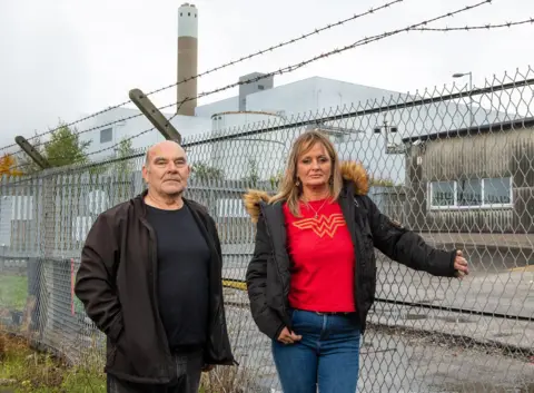 BBC/Jon Parker Los residentes de Lee Runcorn, George Parker, el mecánico y Mandy Royle, se encuentran solemnemente frente a una valla. Al fondo se ve la incineradora de residuos de Runcorn, que aprovecha sus propiedades.   