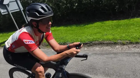 Laura is pictured on her road bike. She has a red and white jersey on and black glasses and a helmet. 