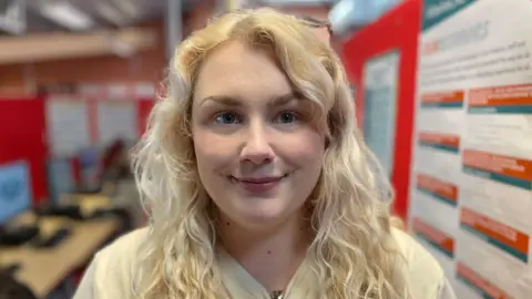 Laura Foster/BBC Charlotte porte ses cheveux blonds bouclés détachés avec un haut blanc. Elle se tient dans une pièce avec des écrans d’ordinateur et des tableaux rouges.