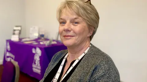 A woman wearing grey jacket, black shirt and white lanyard. She is smiling at the camera and is front of a cream wall, with a table and purple table cloth in the background with leaflets on top