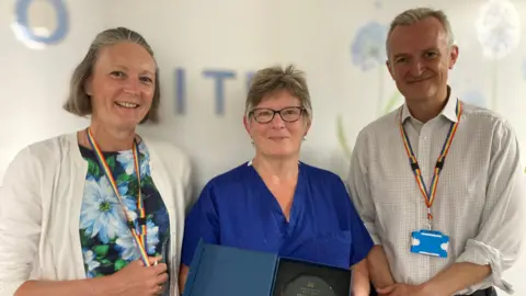 supplied Diane Hele stands in the middle of two colleagues, she is wearing blue scrubs and holding up a glass trophy. Her colleagues beside her (a man and a woman) are smiling at the camera and both wearing lanyards. 