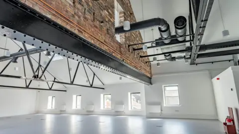 Tom Kay Photographic The top floor of the refurbished Carlton in Maryport. The empty room has white floors, walls and a high ceiling, with a steel beam supporting an original section of the brick wall, from about head height to the ceiling. There are five windows letting light into the room.
