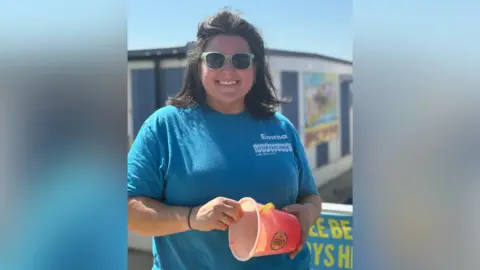 A woman wearing a blue tshirt and sunglasses and holding a small pink bucket smiling into the camera