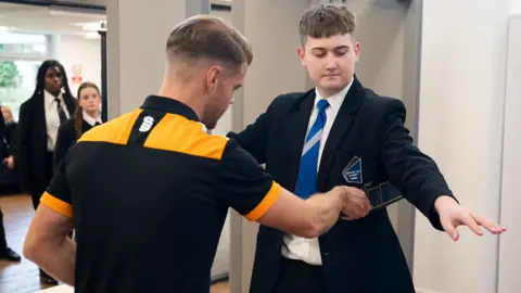 A school boy is scanned with an portable metal detector by a member of school staff. In the background children queue by a doorway. 