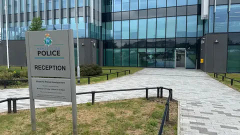 The entrance of a glass and grey cladding-panelled multi-storey building with a grey sign reading 'Merseyside Police Reception' 