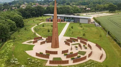 Graham Taylor/IBCC A copper-coloured metal spire in the middle of a huge landscaped memorial garden, which is surrounded by low metal walls in the same colour