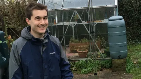 Tim Goodwyn Tim smiles at the camera while standing in his back garden, in front of a glass greenhouse and water butt. He wears a Mountain Warehouse navy and grey anorak. He has dark hair.