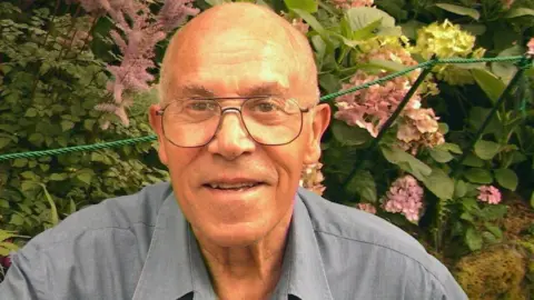 Lucia Dedear An elderly man wearing glasses and a blue shirt. He's sat outside in front of a bush with pink flowers.