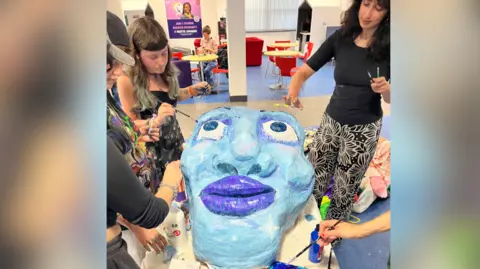 A large papier mache head on a table, a number of women are adding features in different shades of blue. They are all holding brushes. 