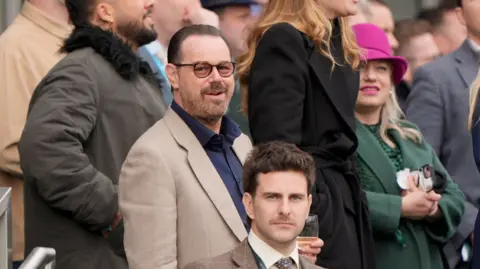 Joe Giddens/PA Actor Danny Dyer is smiling while wearing a brown blazer over a dark blue shirt. He has black-rimmed glasses with orange lenses and is holding a glass. He is standing in a crowd of people.