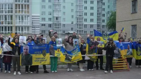 Women wait with blue and yellow flags and posters with pictures of their loved ones