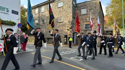 Several men carrying flags march across a road during a remembrance parade in St Peter Port as people and a police officer watch on.