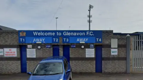 Google Maps A Google Maps screenshot of the exterior of Glenavon Football Club in Lurgan. There is a blue car parked underneath the 'Welcome to Glenavon FC' sign, with four ticket booths within the grey brick wall. There is a metal gate to the right of the image.