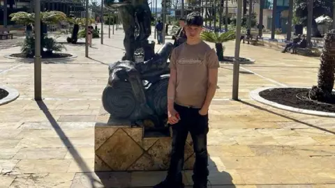 Laura Roberts A young man with short dark hair, wearing a brown t-shirt, standing in front of a statue 