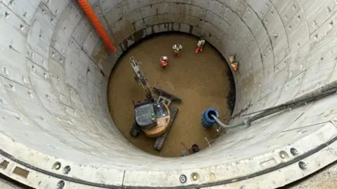 Other An aerial shot of a digger and three workmen at the bottom of a large hole