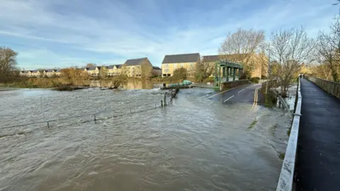 Steve Hubbard/BBC The River Great Ouse has burst its banks and flooded the road. Only the top of the bridge can be seen. Some double yellow lines can also be noticed in the murky brown water. 