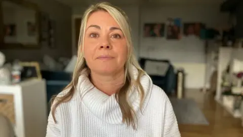 BBC/Seb Cheer A woman, Alison, sits in a living room. There are photos of Livia on a wall behind her.