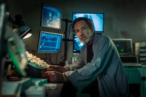 Channel 4 Robert Lindsay, in his role in Generation Z. He is wearing a grey dressing gown and had a bank of computers and screens behind him.