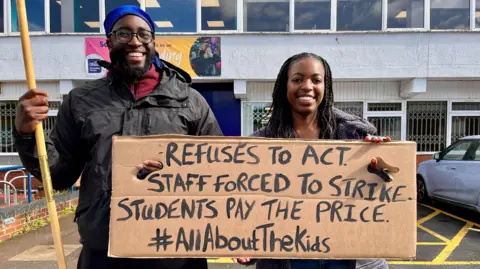 Two people hold a handmade placard. It says "refuses to act, staff forced to strike, students pay the price"