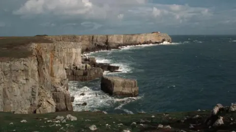 Getty Images Cliffs at St Govans Head. The sky is blue and cloudy. The waves crash into the cliff face.